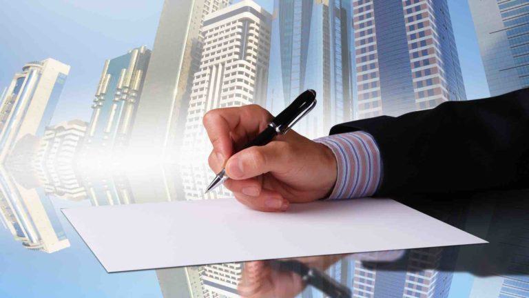 Hand mit Stift setzt zur Unterschrift an, im Hintergrund Hochhäuser